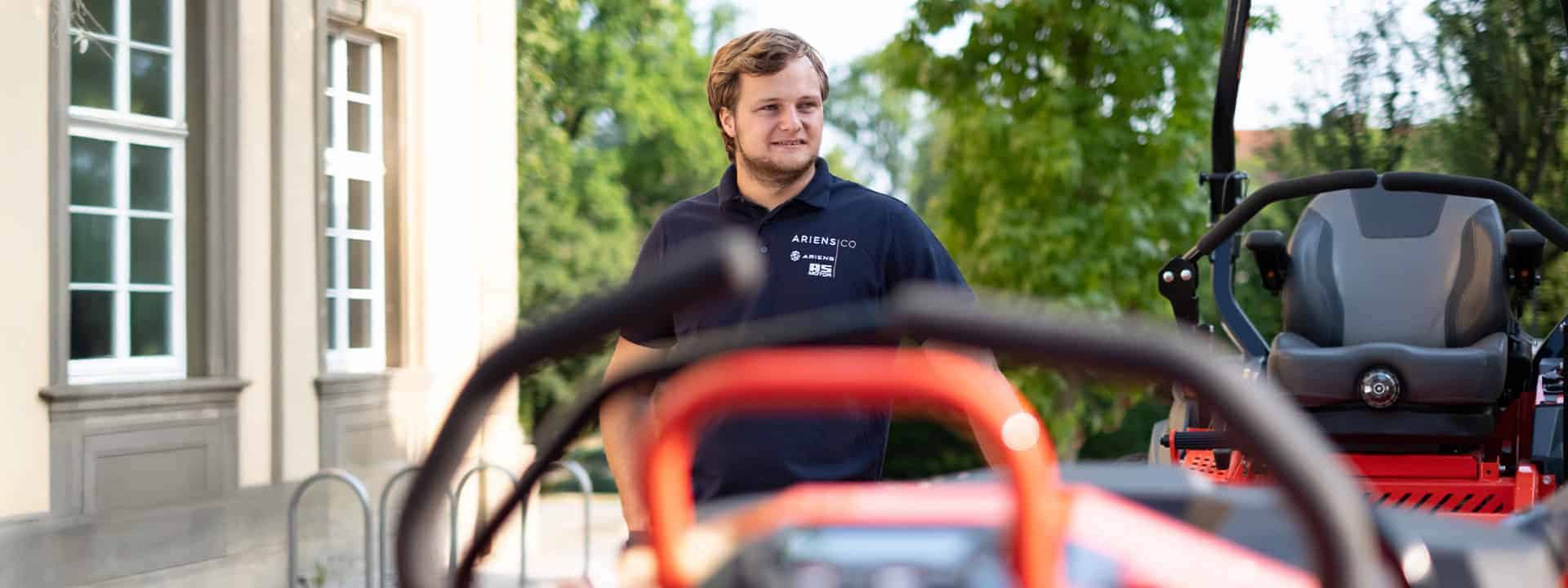Out of focus shot of the control/driver levers of a Zero-Turn mower in the foreground and in the background a standing smiling man